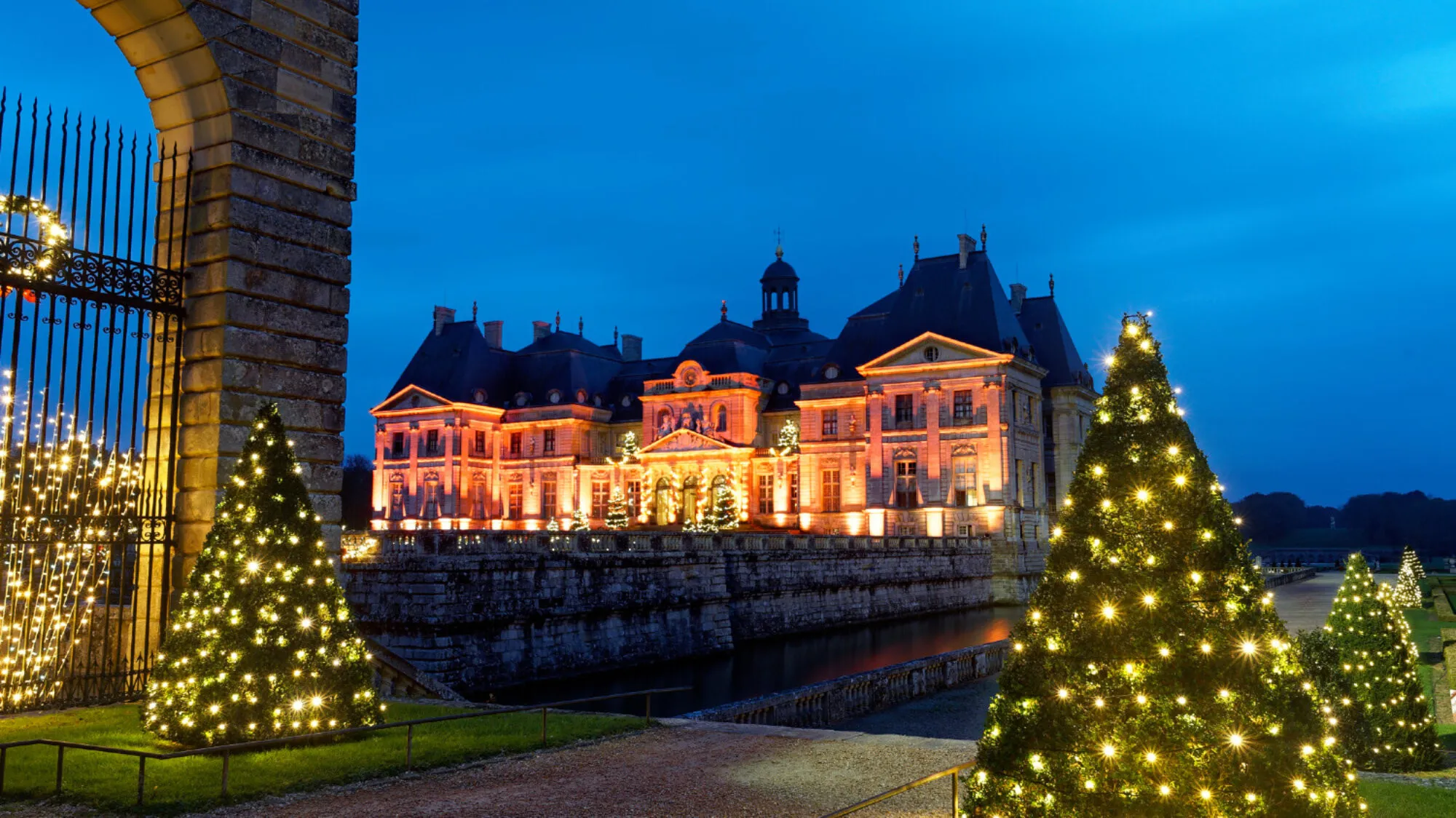 Arrival at Vaux-le-Vicomte Christmas Evening Vaux-le-Vicomte château at dusk framed by a stone gate and cone-shaped Christmas trees covered in twinkling lights