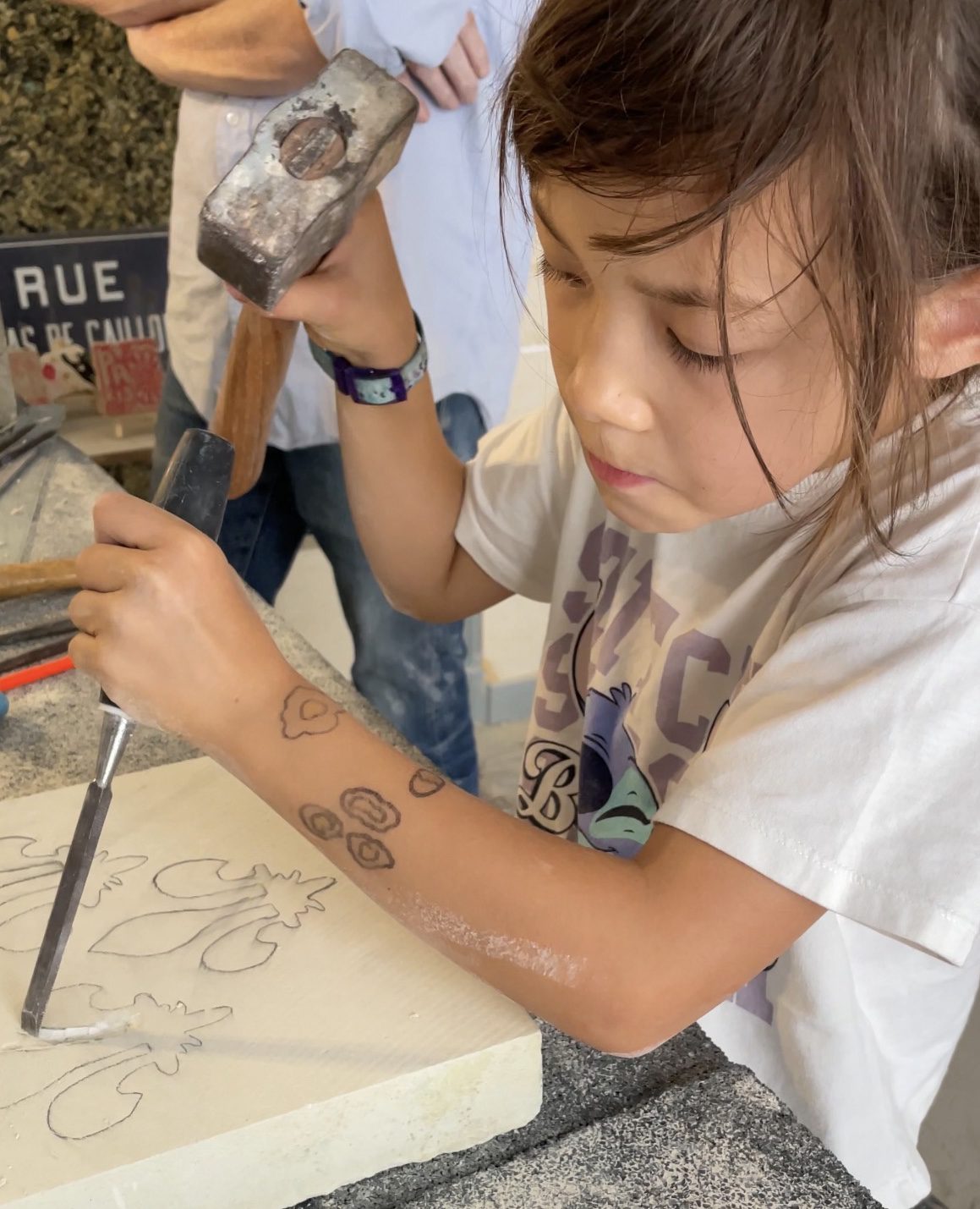 Kid-Friendly Stone Cutting Workshop in Paris Young girl concentrating as she uses a hammer and chisel to carve a fleur-de-lis design into a stone block during a family workshop