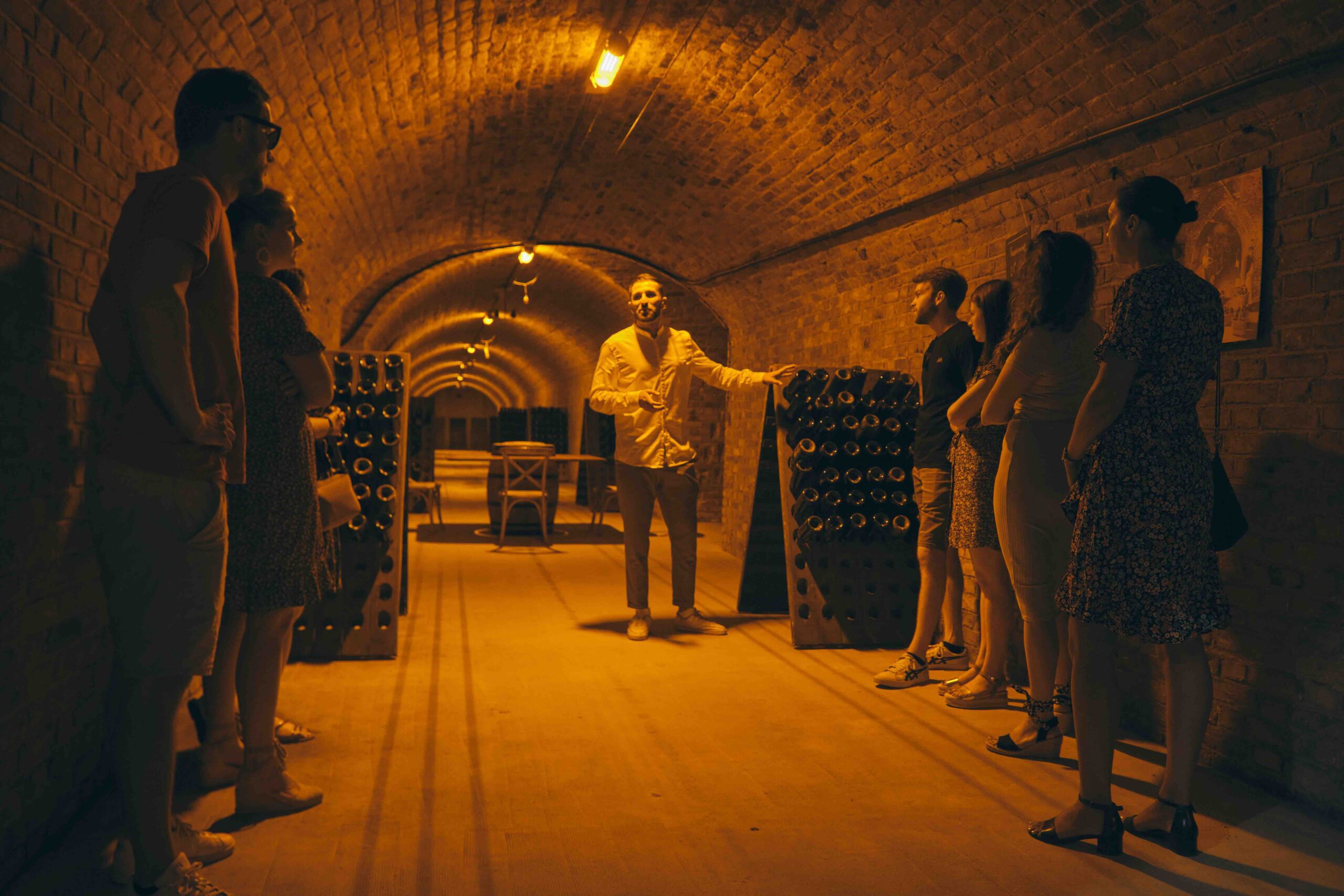 Champagne cellar stop on day trip by train Small group stands with a guide in a warm-lit Champagne cellar, listening to explanations surrounded by racks of aging bottles.