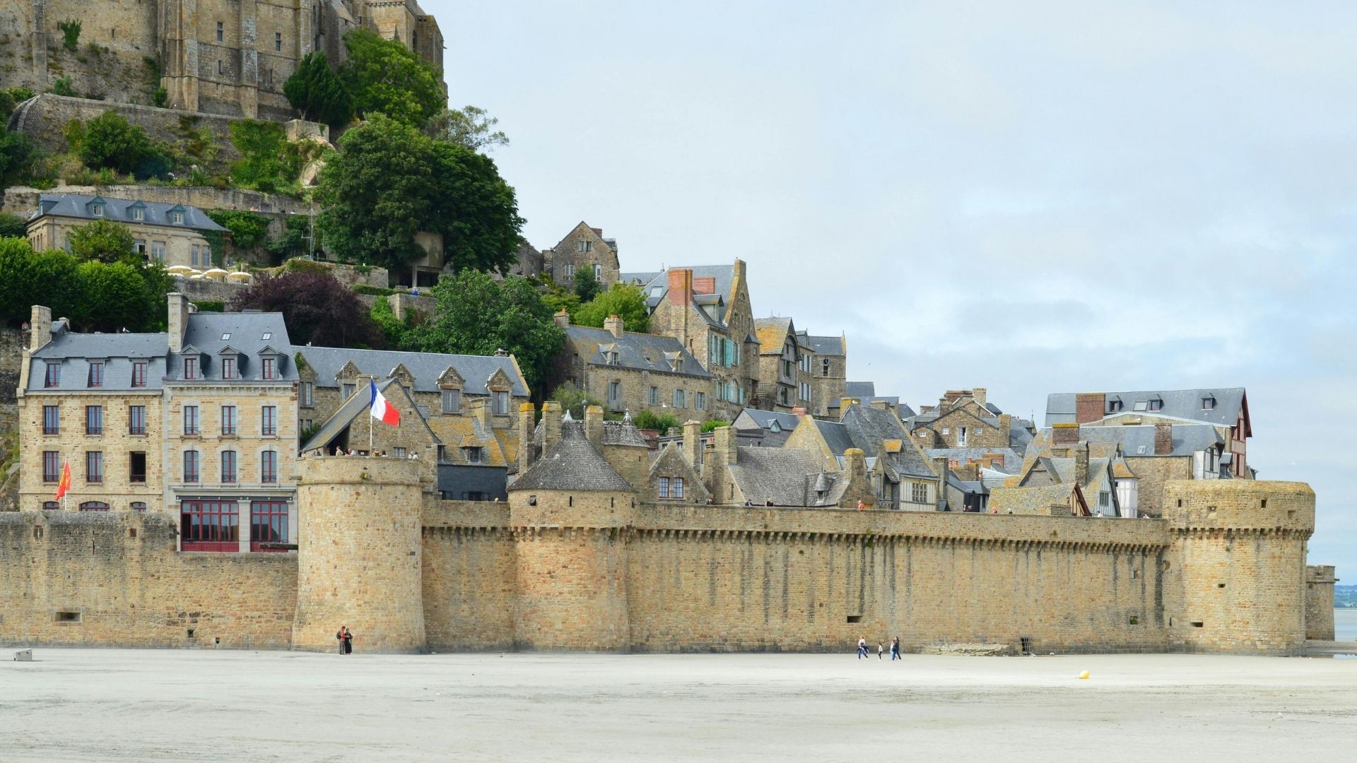 Mont Saint-Michel Ramparts Low Tide Paris and Beyond Tours Village Architecture Experience Mont Saint-Michel medieval rampart walls and village houses cascading up hillside with French flag during low tide - Paris and Beyond Tours private guided tour from Bayeux Caen